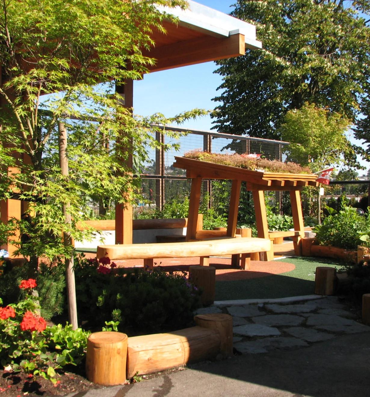 Timber pergola with green roof and log benches in a garden play area beside red flowers