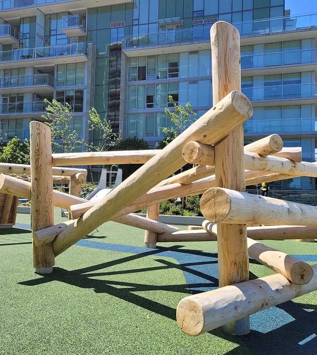 Peeled-log climbing structure on green rubber surfacing in front of a glass apartment building