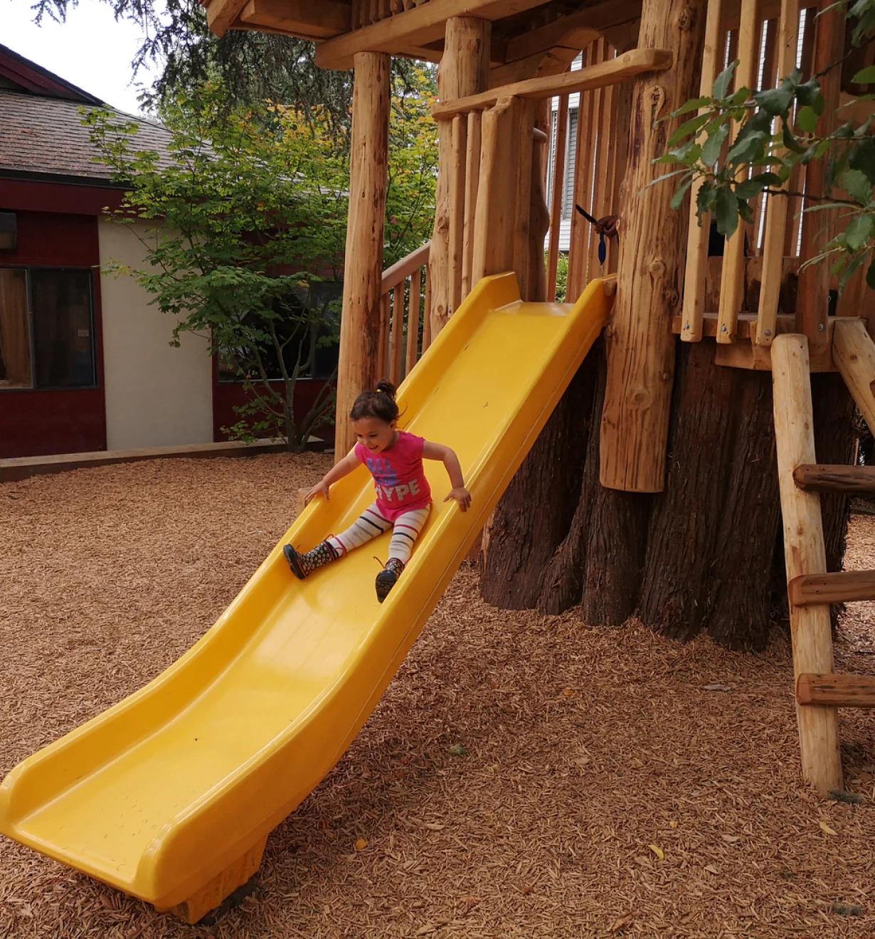 Child sliding down a yellow slide from a log play structure on a wood-chip playground