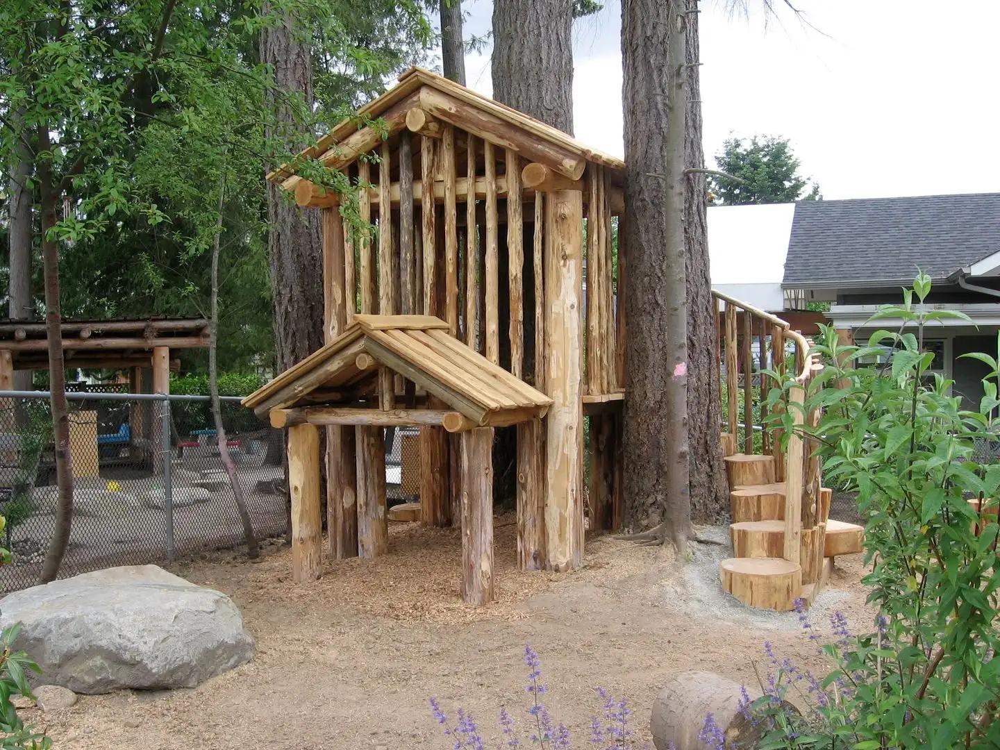 Log cabin playhouse with a covered porch, stump steps and a boulder on sandy ground